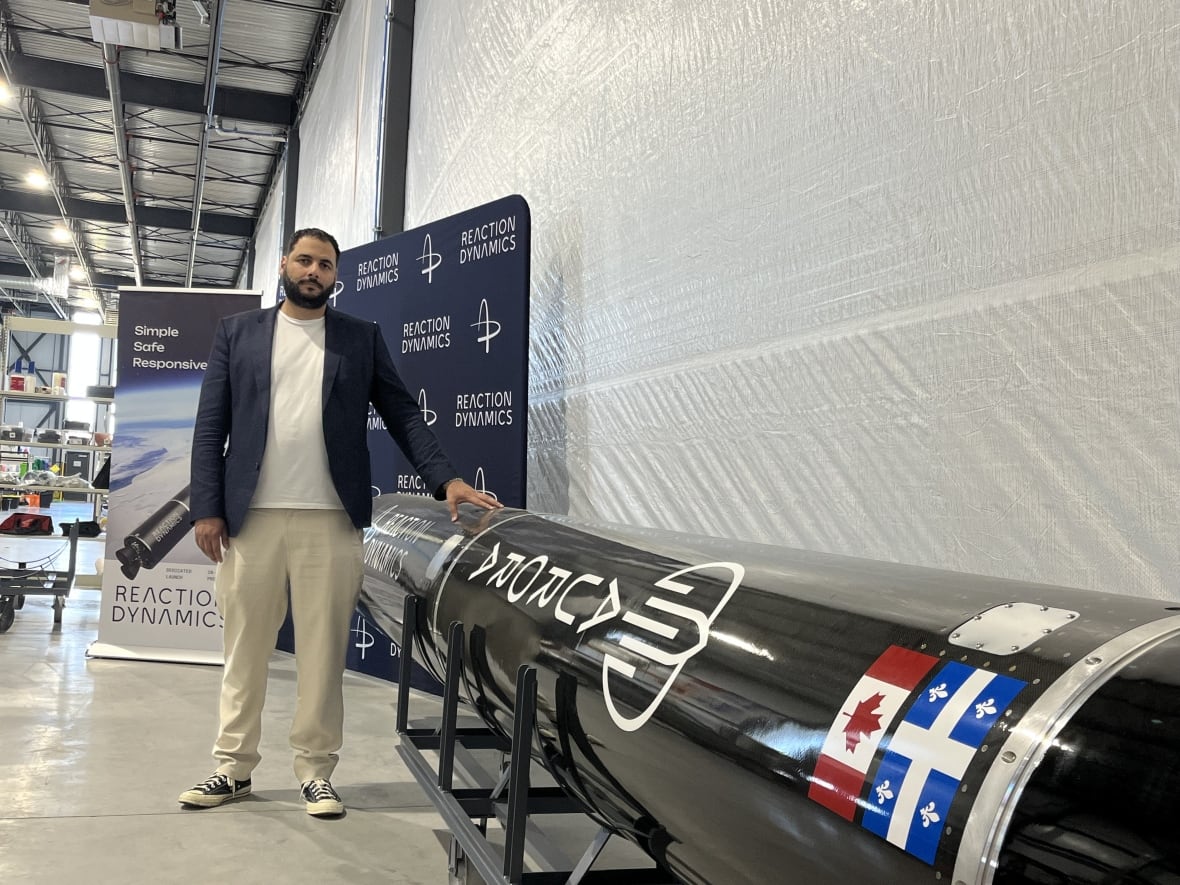 A man in a white shirt, a dark-blue jacket and khaki pants, stands with his hand on a black rocket.