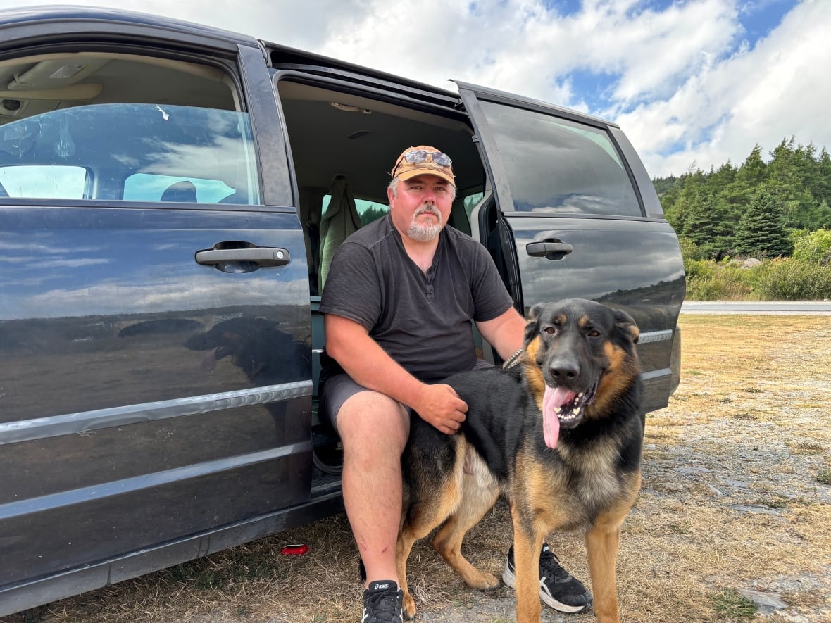 A man sits on the side of a mini van. He is holding onto a large dog.