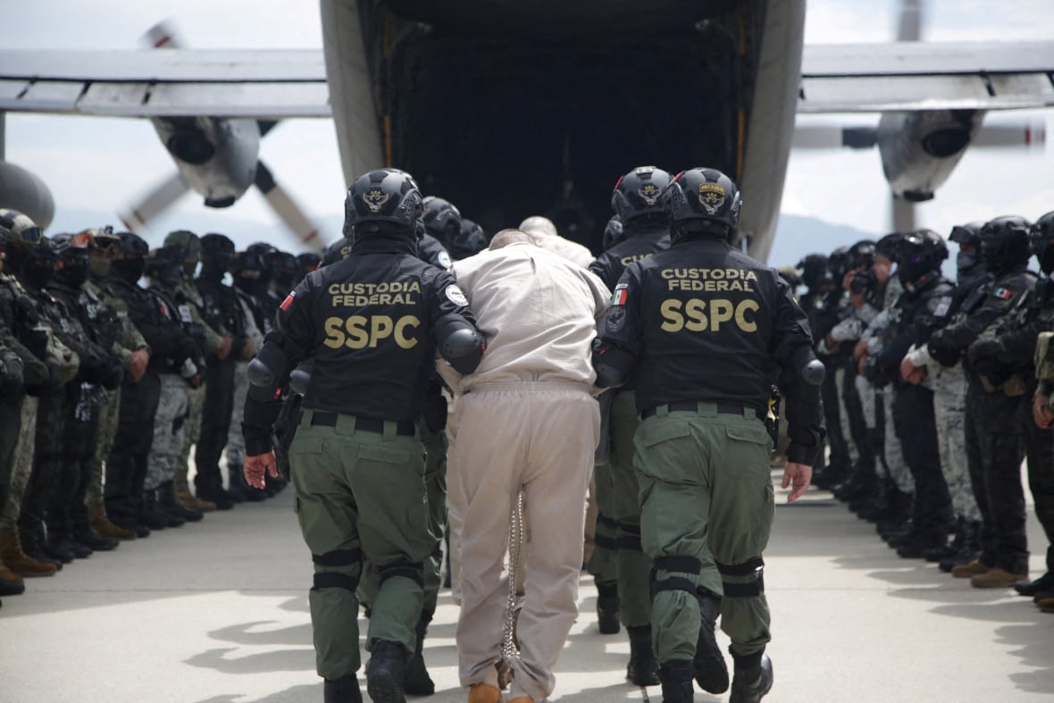 A handcuffed man in beige jumpsuit being frog marched by federal police towards a plane that's on a tarmac and flanked by other officers.