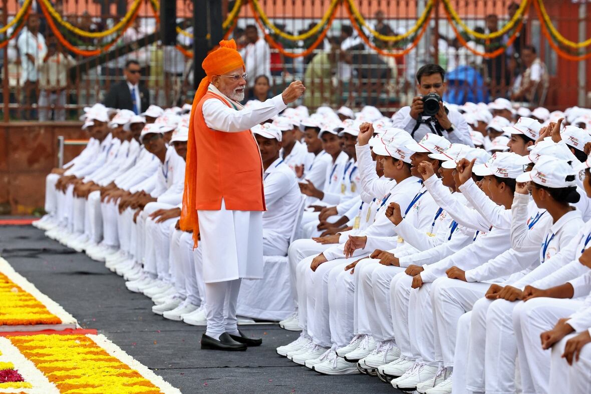Indian Prime Minister Narendra Modi, who wears white and bright orange, stands in front of a group of young white with their grip in the air. 