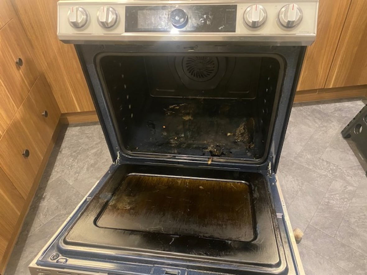 In a kitchen with wood panel siding, an oven with a silver colour control panel and round control knobs has the door open and has burned black material showing on the inside.