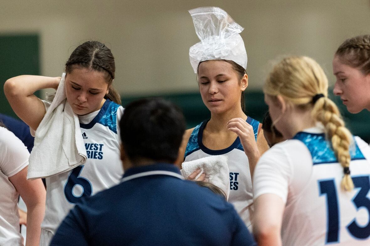 A huddle of female basketball players, one wiping the back of her neck with a towel, the other with a plastic bag of ice on her forehead.