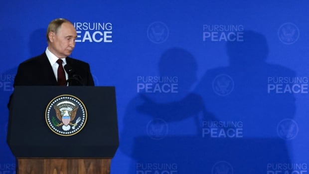 A man standing at a podium during a news conference looking downward and casting a shadow on the backdrop that reads Pursuing Peace. next to the shadow of another man.