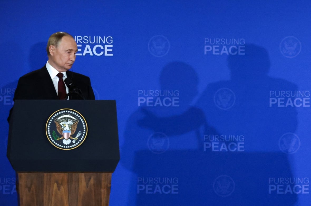 A man standing at a podium during a news conference looking downward and casting a shadow on the backdrop that reads Pursuing Peace. next to the shadow of another man.