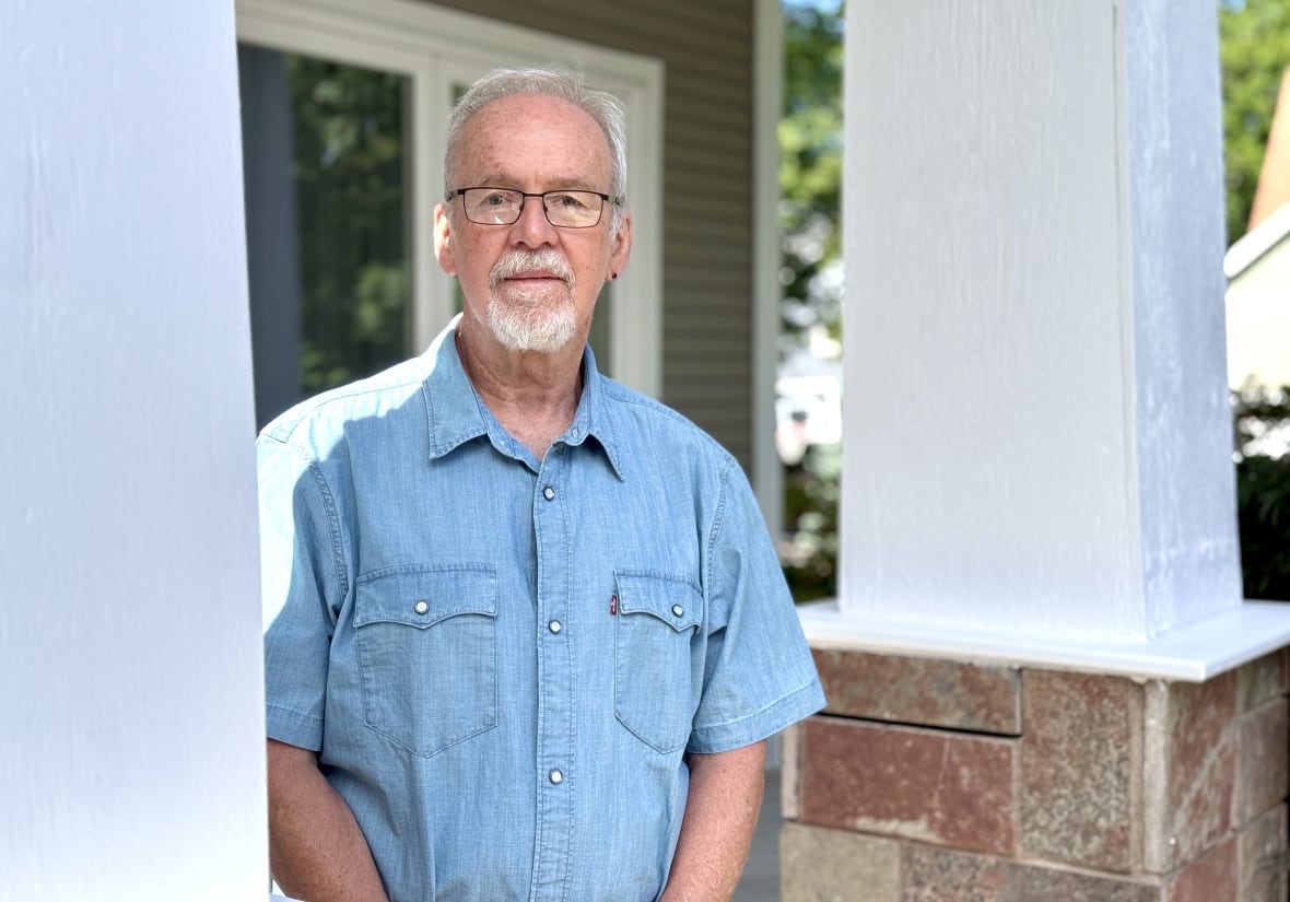 A man wearing a blue short sleeved collared shirt stands outside on the entryway of a house.