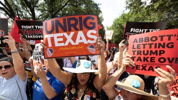 Several people, men and women, hold signs in an outdoor demonstration. A woman in a white hat near the front holds a sign over her head which reads, 