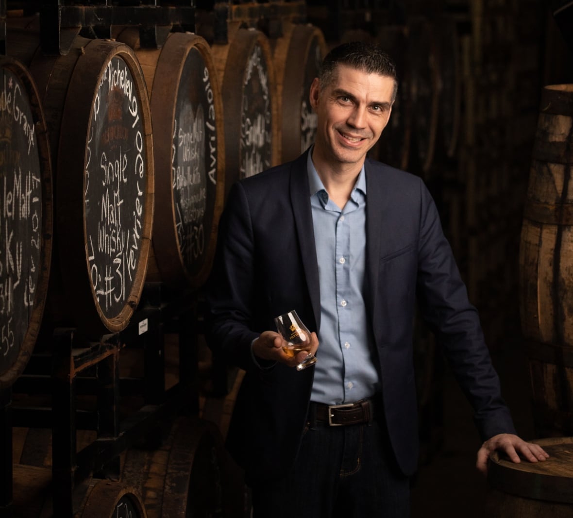 Ein Mann in einem schwarzen Blazer und einem blauen Hemd mit Kragen darunter hält ein kleines Glas vor Fässern in einer Brennerei.