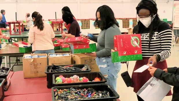 Young volunteers in face masks work in a large gymnasium filling red and green boxes, drawing from bins filled with small toys and items on tables before them.
