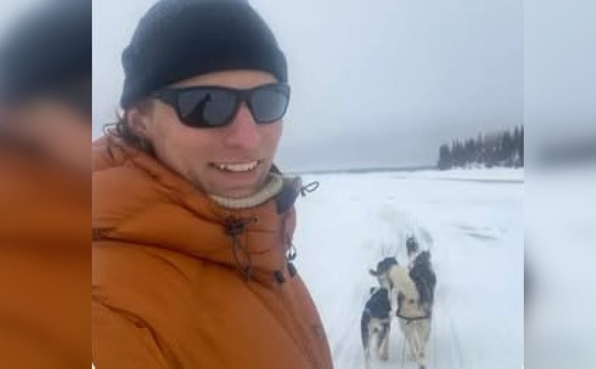 A man wearing black tuk, orange bars and black glasses heading towards the camera. It is snowy outside. There are dogs in the background.