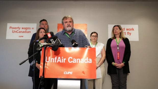 Supporters stand behind a union leader who is speaking to the media from a podium that has a sign reading: UnfAir Canada, as well as signs posted on the wall behind them including one that reads: Poverty wages = unCanadian.
