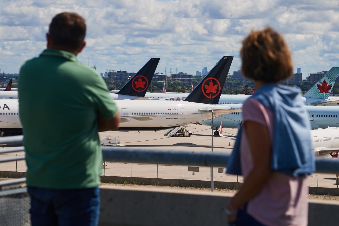 Standing outside an airport, two travellers look at grounded Air Canada planes on the tarmac.
