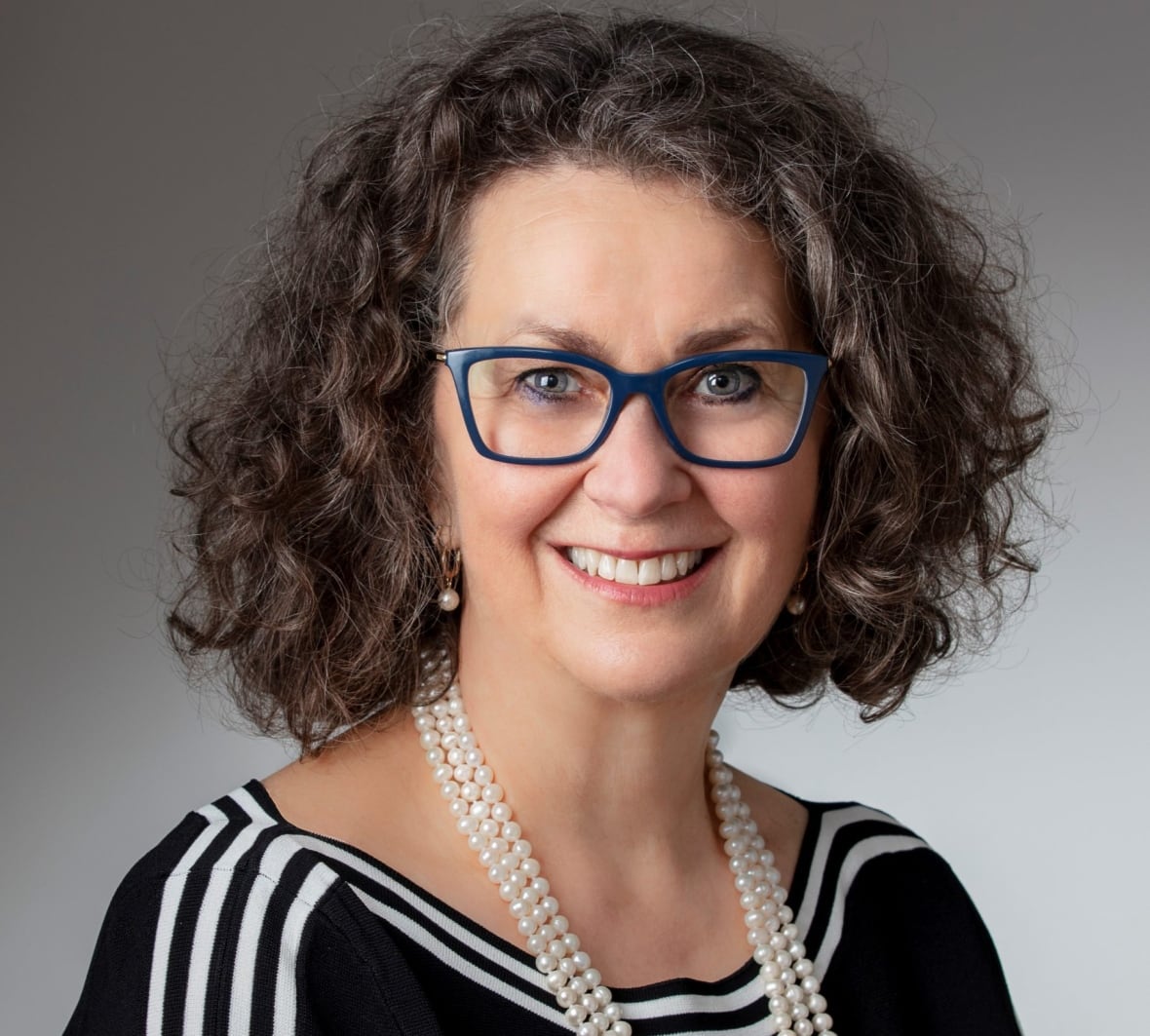 Eine Frau in schwarzer Bluse, weißer Perlenkette und blauer Brille lächelt in die Kamera.