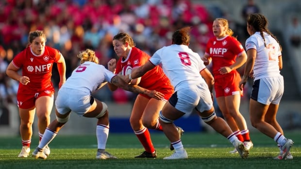 A female rugby player tries to run with the ball through two defenders.