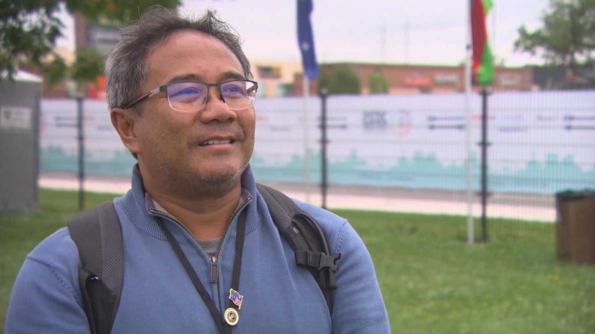 A man in a light blue sweater and glasses smiles while being interviewed by a reporter.