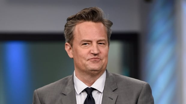 A man in a suit appears in front of a blurred studio background. 