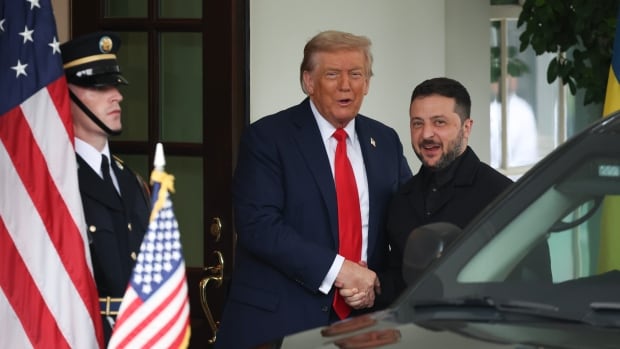 An older cleanshaven blonde haired man and a younger bearded dark haired man shake hands in front of a building in an outdoor photo. Near them, a guard stands near the American flag of stars and stripes.