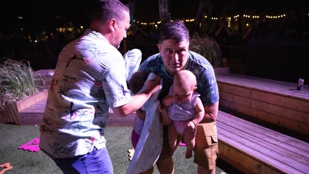 Roberto Mendoza holds his daughters as his partner, Jean-Philippe Forget, shields their eldest with a blanket, in a widely shared photo taken Aug. 9. A man holding a baby, a young girl clings to him as another man shields her face with a blanket, standing on a patch of fake grass with kids toys