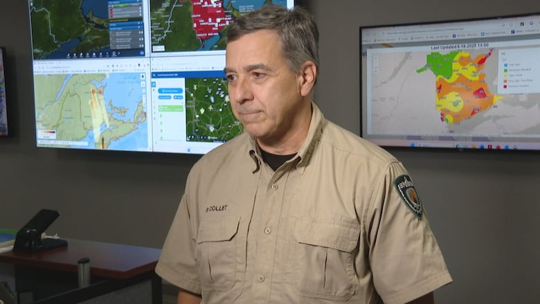 A man in a beige shirt standing in front of TV screens.