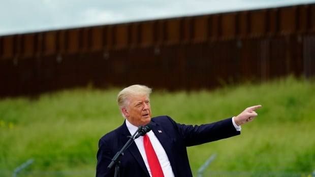 A cleanshaven older man in suit and tie gestures, near a large wall-like structure outdoors.