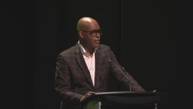 A bald man with glasses and a dark plaid suit stands at a podium addressing a crowd