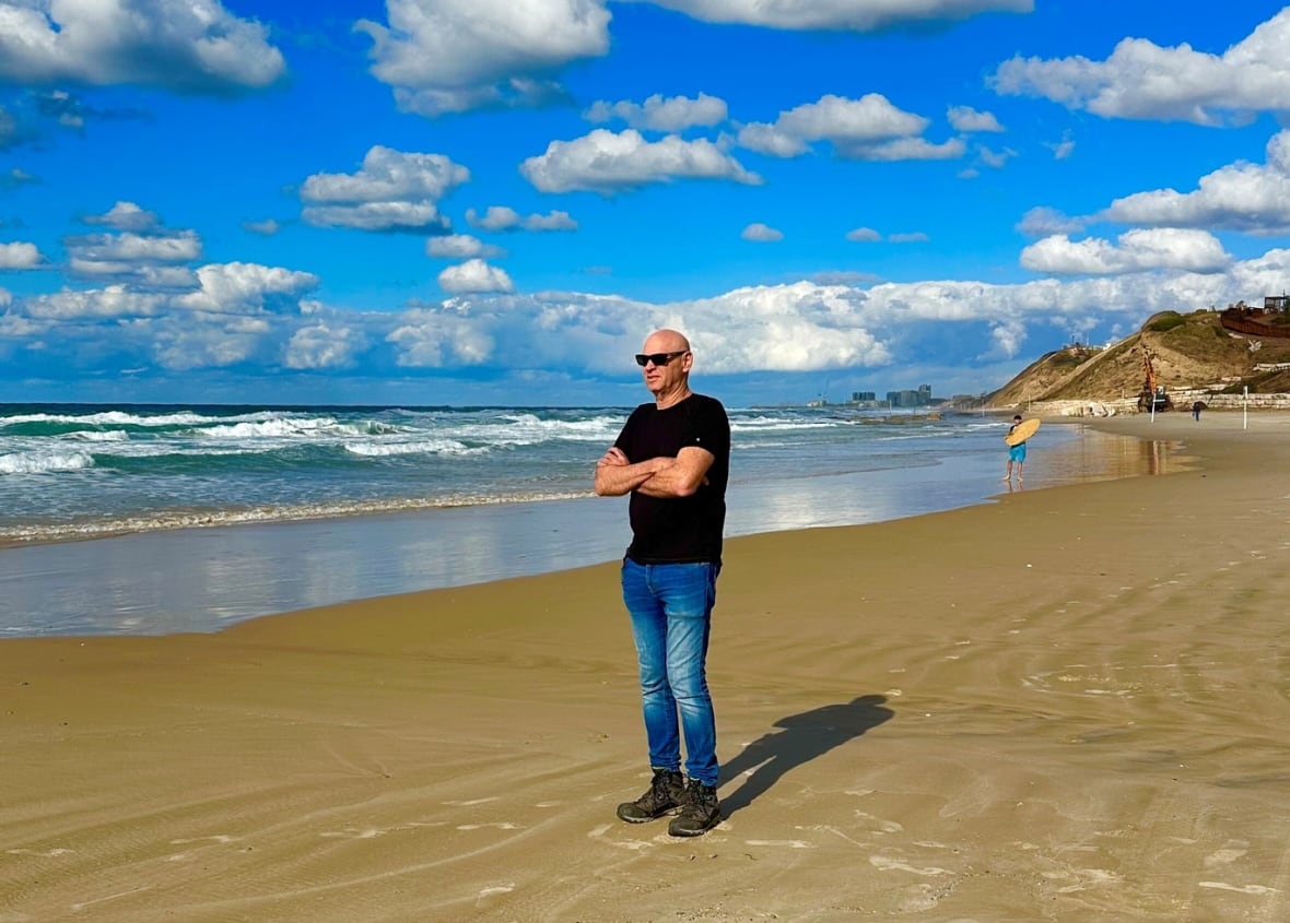 a man in jeans, a dark tshirt and sunglasses stands alone with his arms crossed on the sand of a beach. the sky is blue overhead and filled with white, puffy clouds.