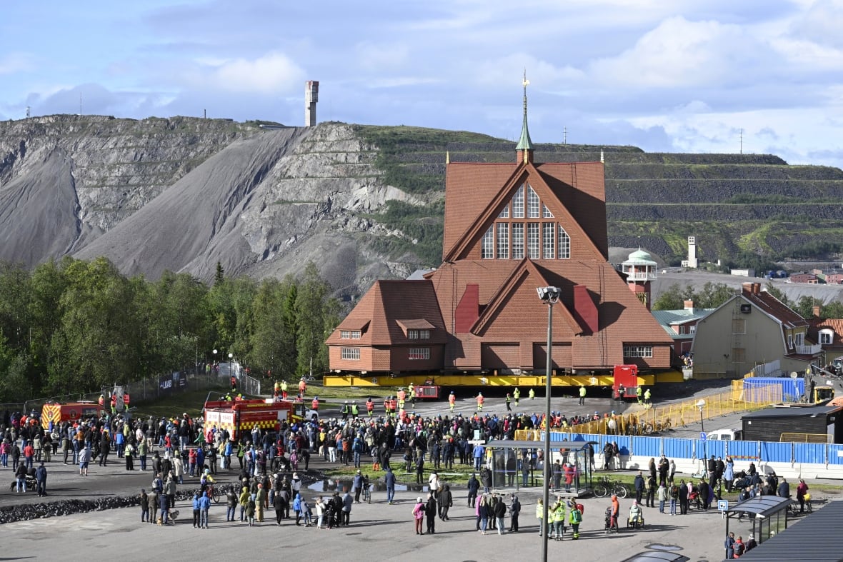 A crowd of people stand outside in front of a large, brown, wooden A-frame church hoisted on yellow beams, with mountains on the horizon.