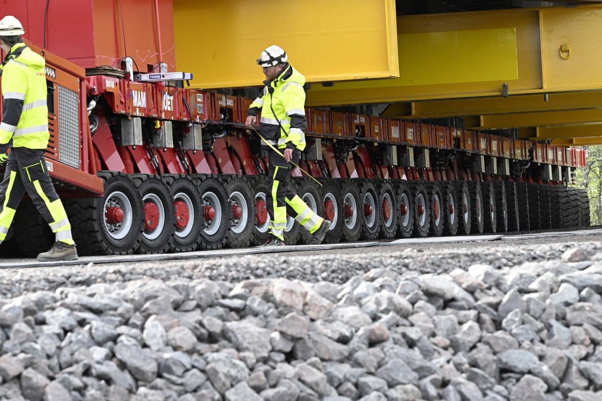 People in bright yellow safety suits walk alongside dozens of wheels. 
