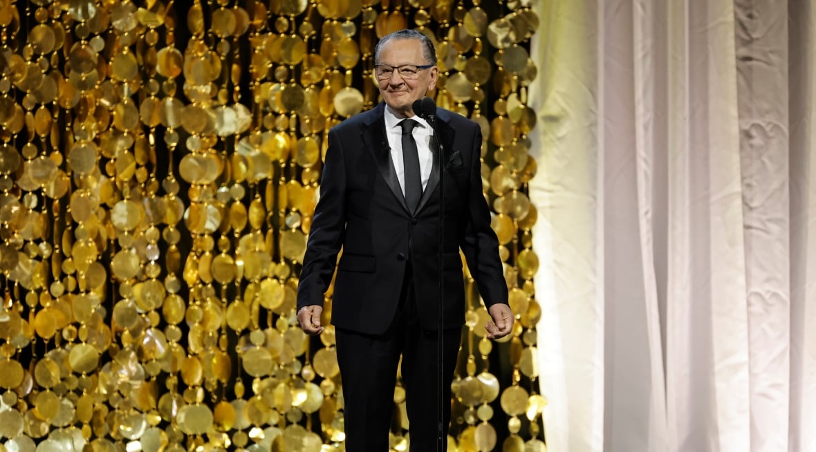 A man wearing a suit on the stage stands in front of the glossy gold wall. 