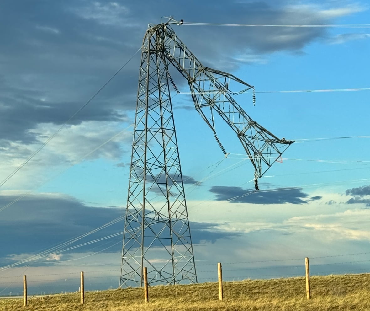 A power transmission tower is bent downwards, with the top half hanging down. The sky is blue with dark clouds seen.