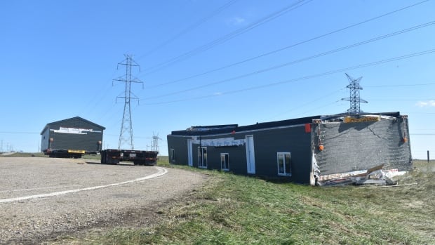 A modular home is seen flipped over upside down. An empty wheeled flatbed is beside it. In the background, another modular home remains upright on a wheeled flatboard.