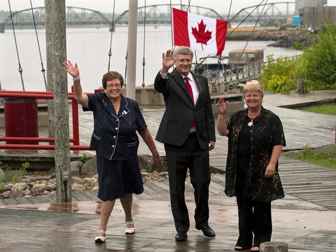 Two women and a man walk on a waterfront boardwalk with a Canadian flag in the background. 