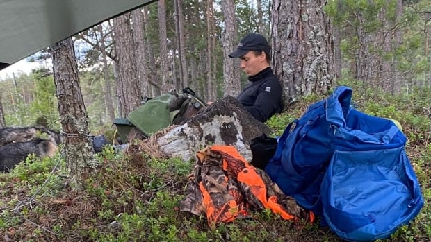 A man sits on a shrubs under a tarp next to outdoor equipment.