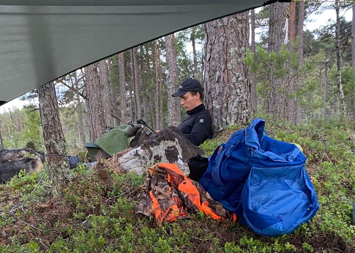 A man sits on a shrubs under a tarp next to outdoor equipment.