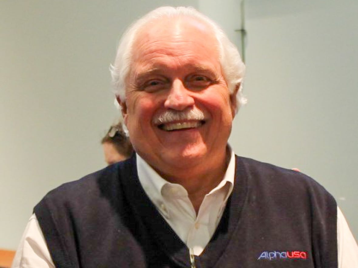 A man with short white hair and a stylish white mustache while wearing a jacket jacket with Alphaplus on a white shirt.