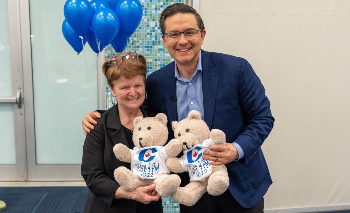 A woman and man smile as they stand in front of blue balloons and hold teddy bears with small T-shirts saying "Pierre for PM 2022."