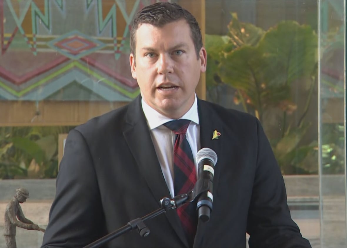 Kody Blois, Parliamentary Secretary to the Prime Minister, wears a black suit and speaks into a microphone. 