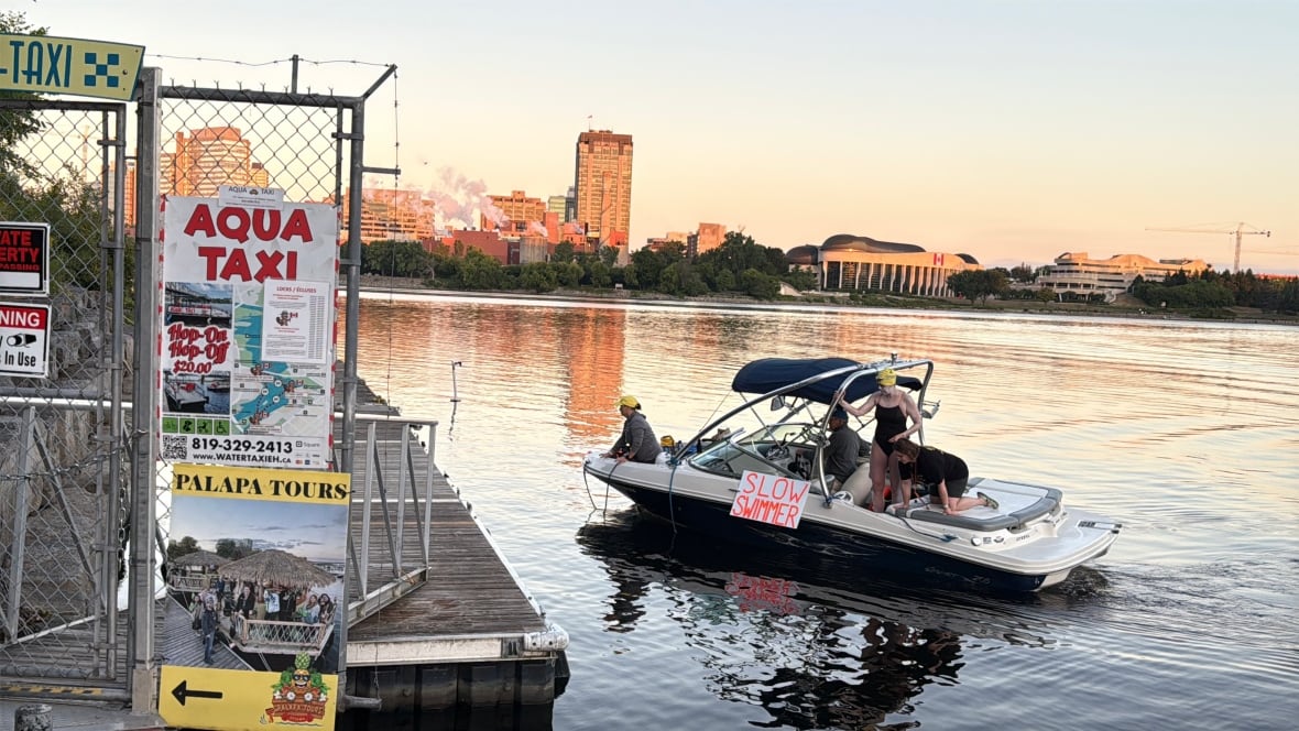 A woman in a black swimsuit stands on a boat with a sign that reads 'SLOW SWIMMER' along the Ottawa River, next to a water taxi stand.