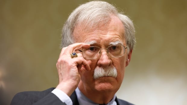 A white haired man with glasses and thick mustache adjusts his glasses in closeup photo.