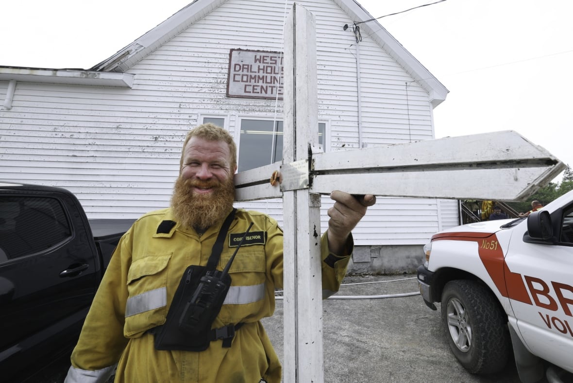 A man in yellow firefighting clothing holds a large white cross in his left hand.