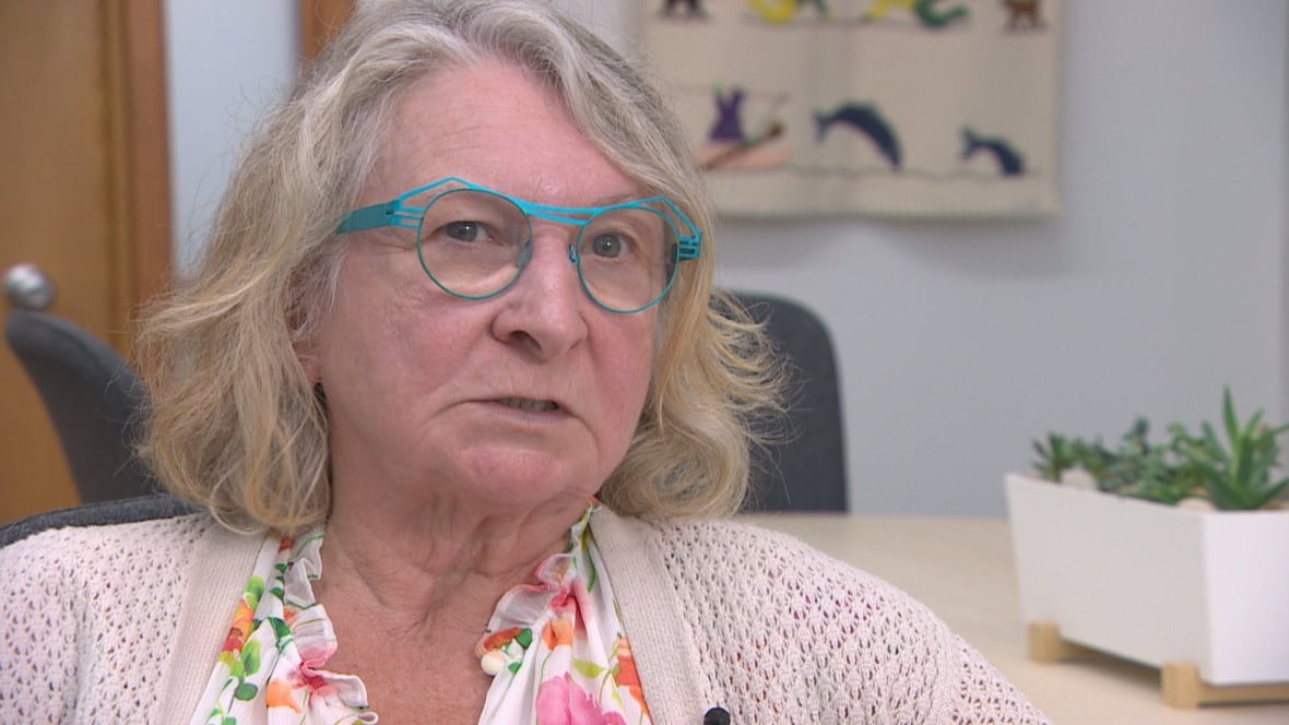 A woman with light, wavy hair, teal-rimmed glasses, a pale pink sweater and floral blouse looks slightly off-camera to the right.