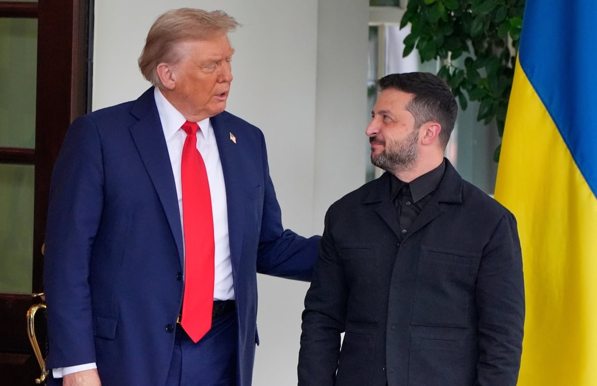A man wearing a blue suit and a red tie touching the arm of a bearded man wearing a black suit.