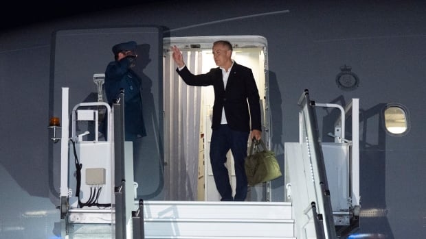 A man in a suit about to board a flight waves from the top steps leading up to the plane.