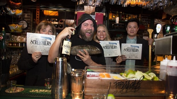 Man in a black hat smiling in a bar, holding a bottle of rum and a cod. People next to him are holding sheets of paper with word 'screechers' on it.