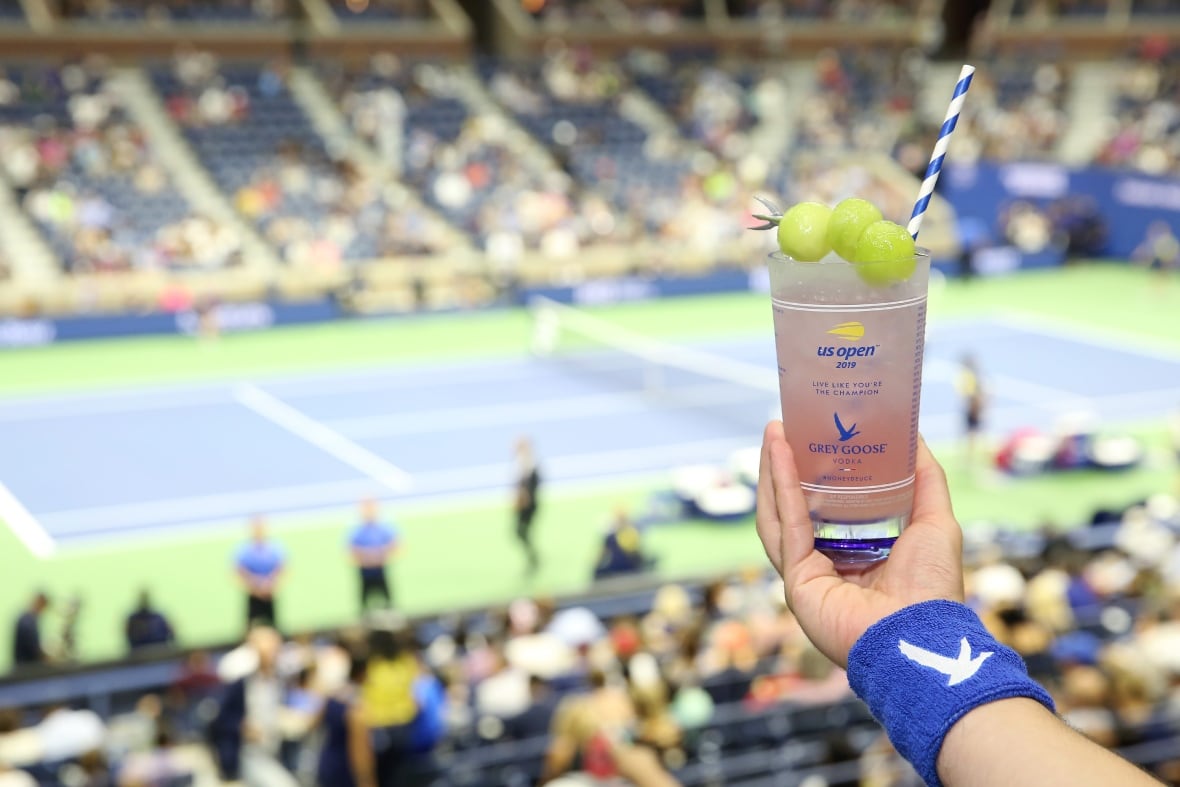 A hand holds a cocktail in front of a tennis match