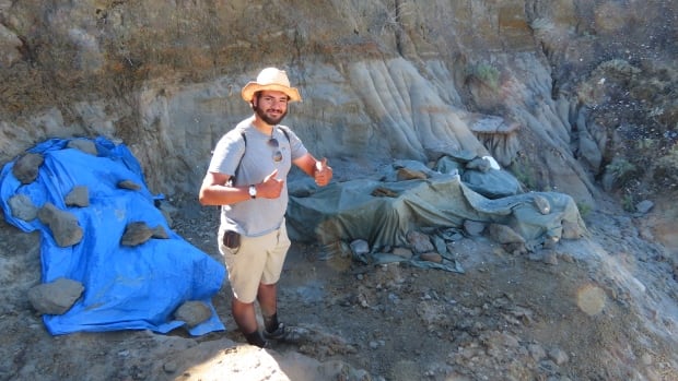 A man with a dark beard and a tan hat stands in front of a rock hill holding his thumbs up.
