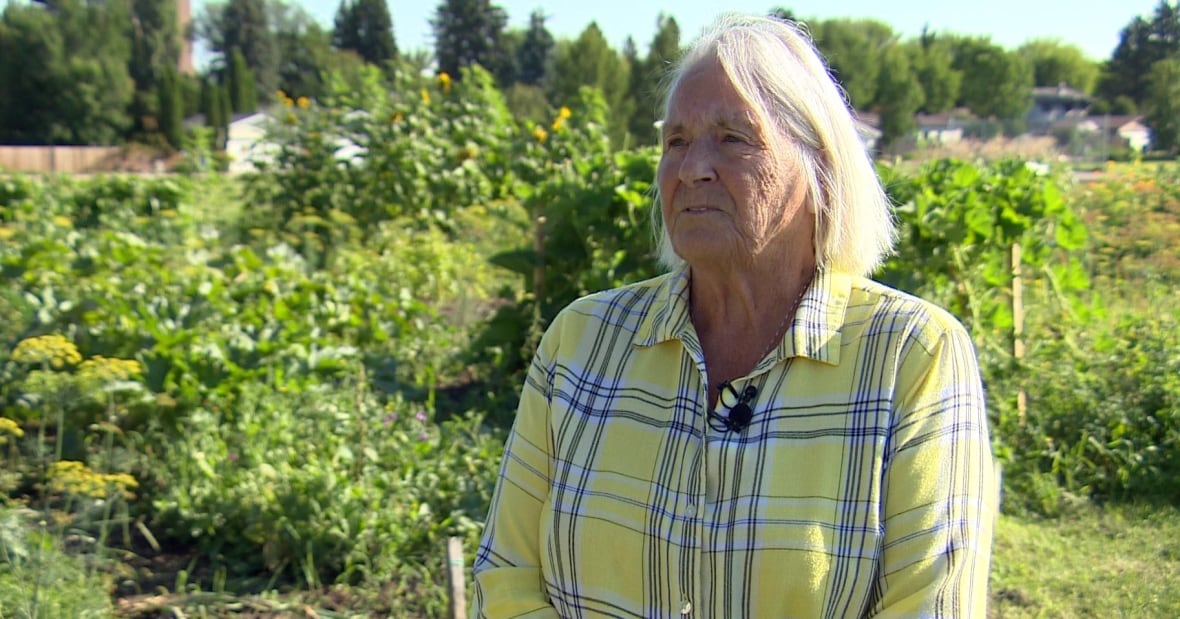 An elderly woman with white hair stands in a garden wearing a yellow shirt. 
