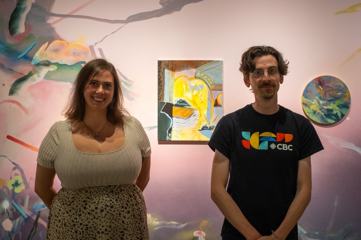 A woman with a light-coloured top, a printed skirt and long hair stands beside a man with medium-length hair, glasses and a goatee. Behind them is a wall covered in colourful abstract paintings. 
