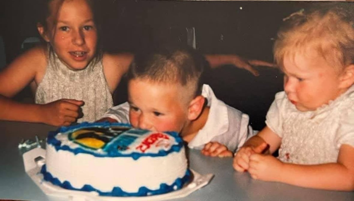 Three kids sit at a table while one leans into a birthday cake.