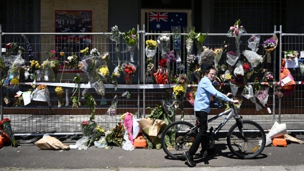 Several wreaths and bouquets of flowers are shown laid out on a sidewalk near a building. A male bicyclist rides by. 