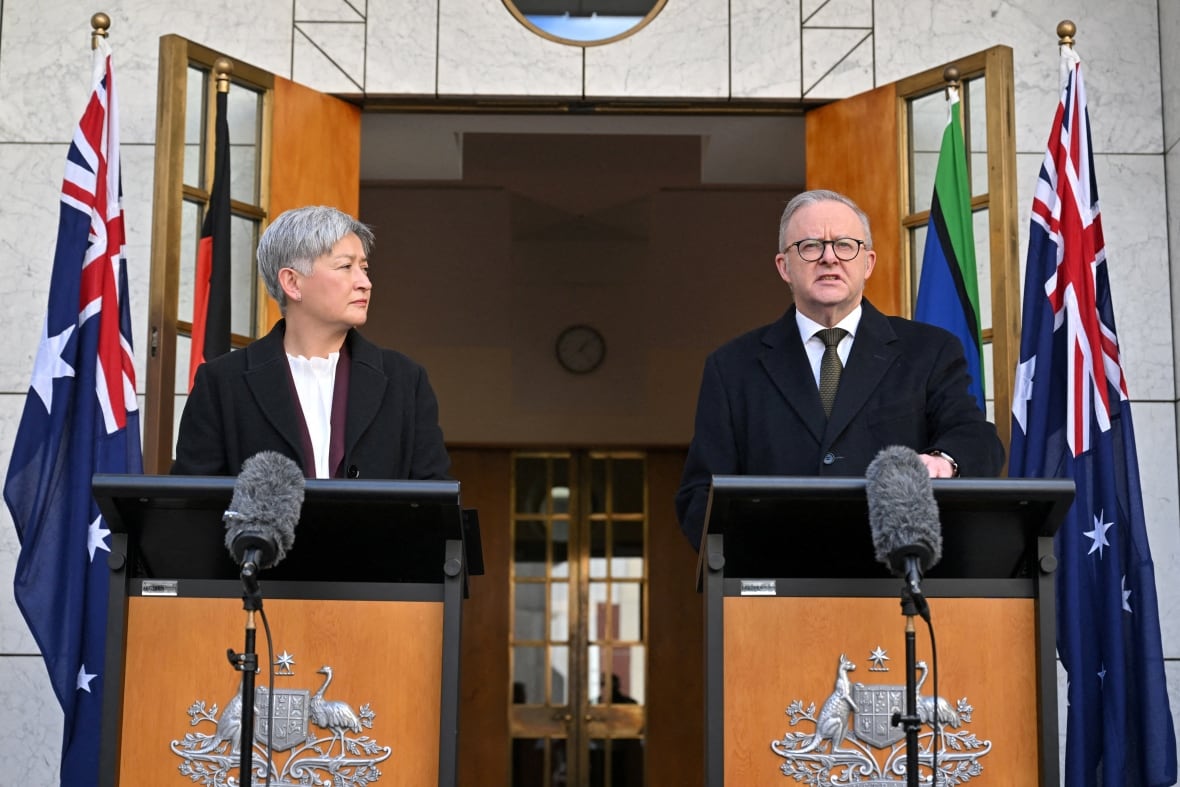 A clean man and a tangible man speaks in a suit and tie on a platform in front of the flags while a lyric woman searches on a separate platform.